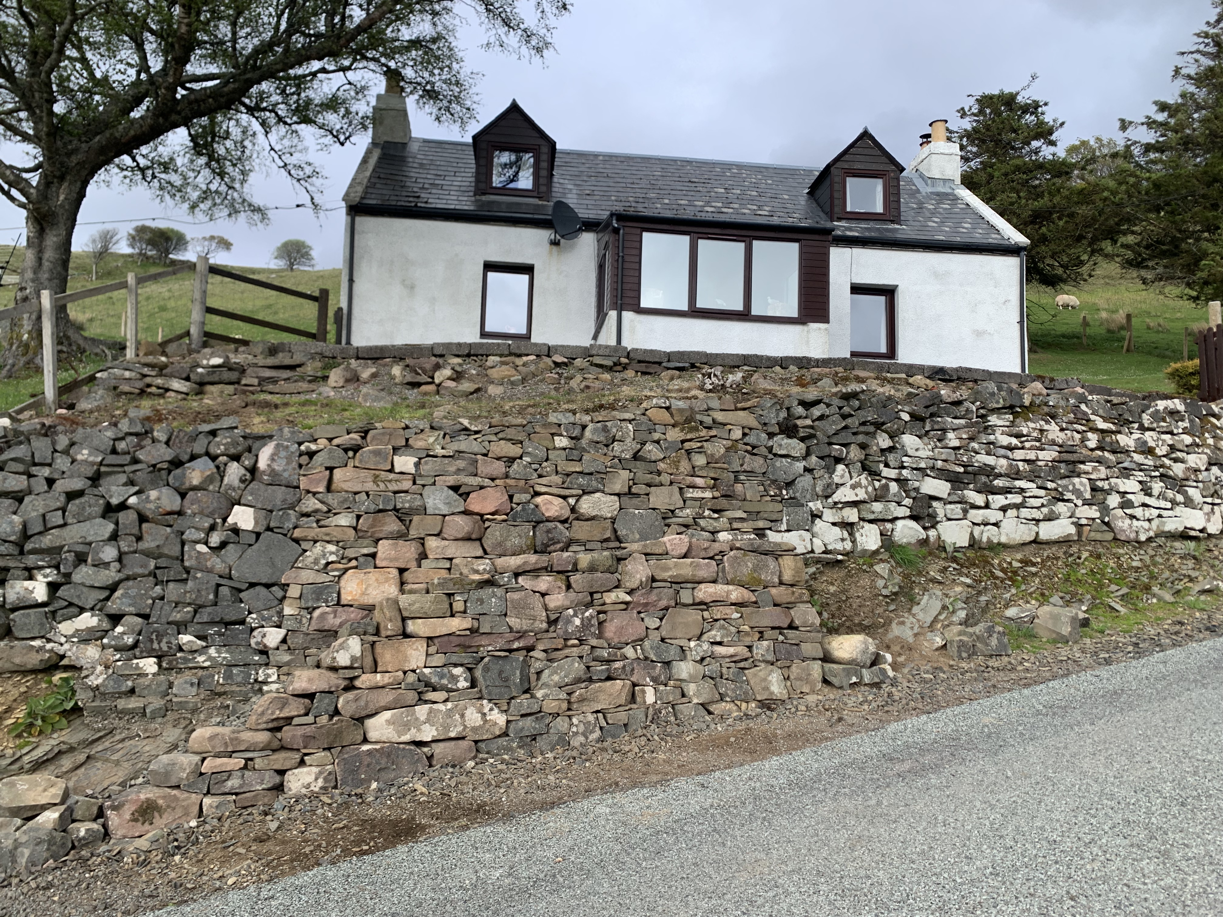 A high dry stone retaining wall with the central section repaired, a cottage above.