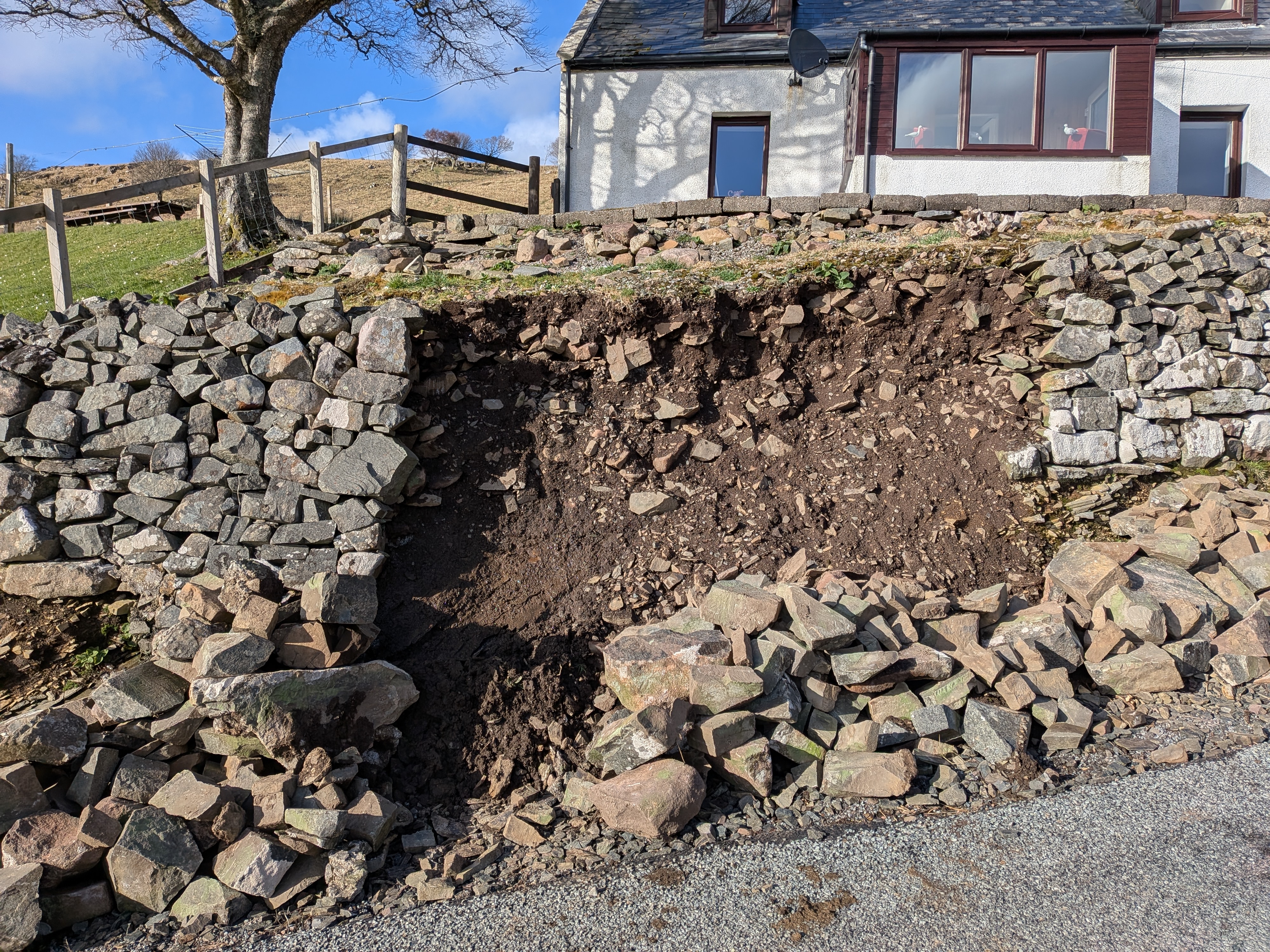 High retaining wall with the central section having collapsed revealing an earth bank, a cottage above.