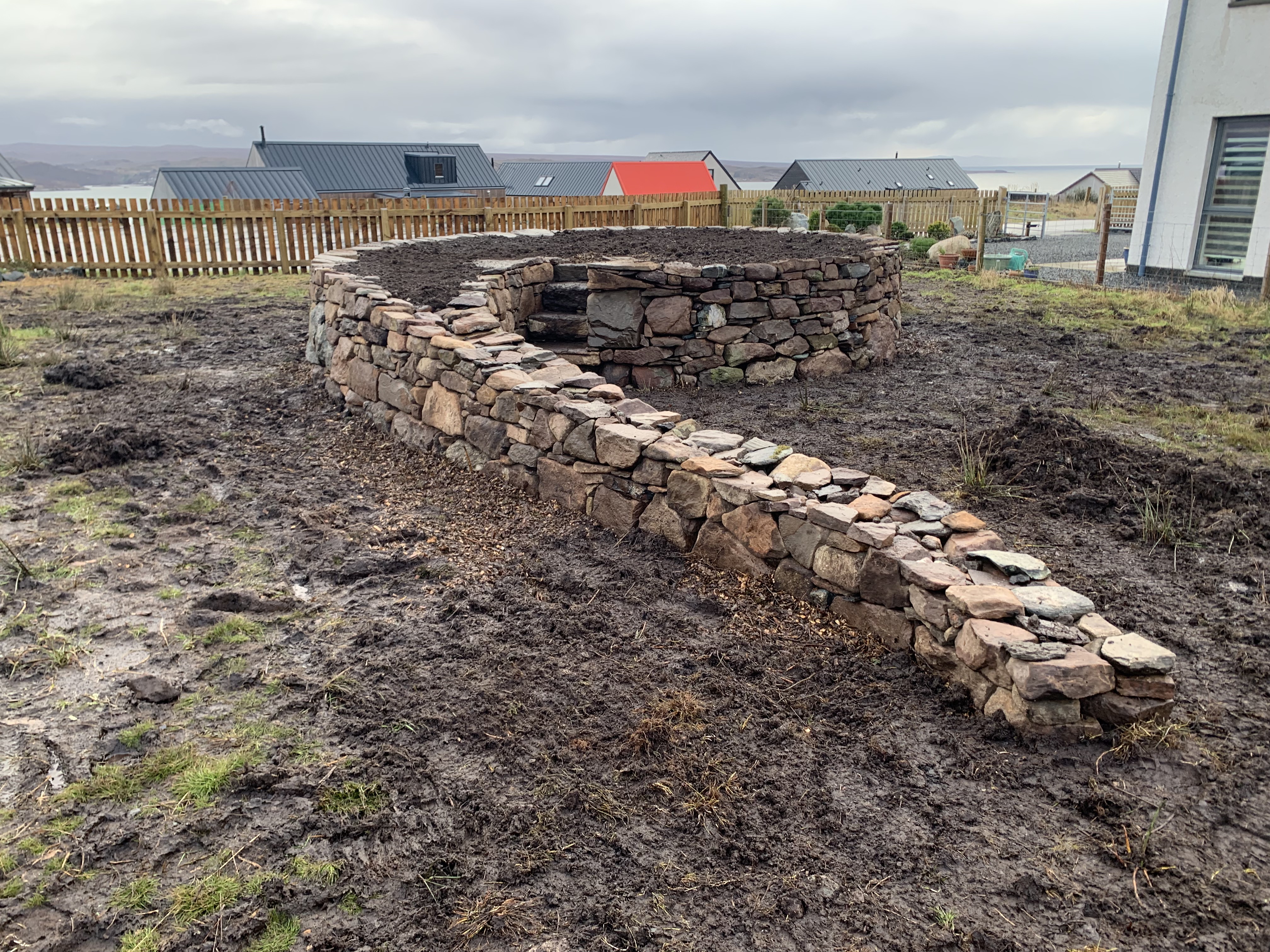 A circular dry stone raised bed structure with a curving tail in the foreground, like a tadpole, 