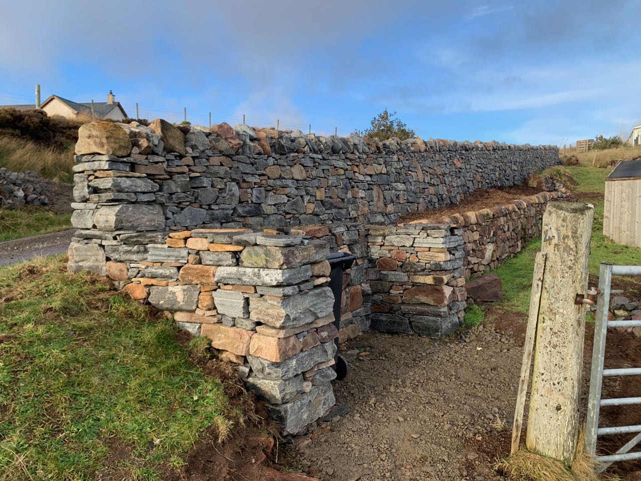 A newly built dry stone wall running up a hill with a dry stone bin store in the foreground.