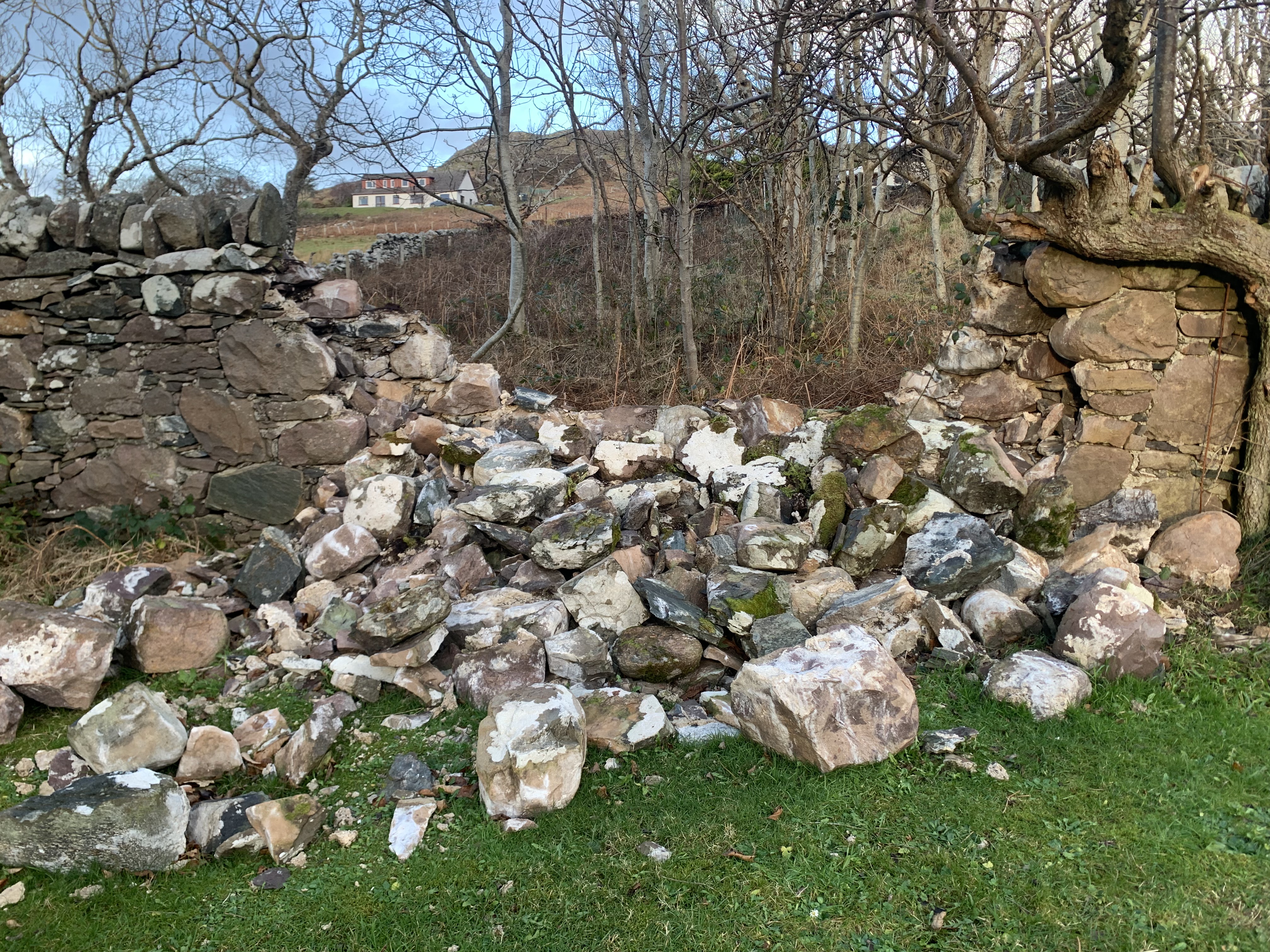 A disorderly tumble or large stones forming a gap in a dry stone wall with trees in the background