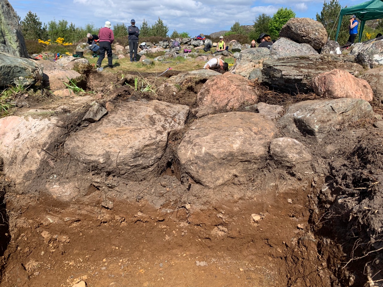 Large foundation stones of a roundhouse exposed during an archaeological dig, museum volunteers in the background